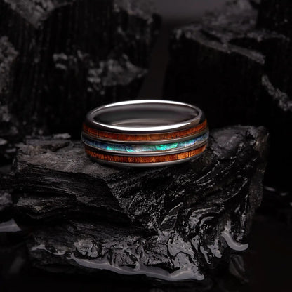 Brushed silver tungsten ring with abalone shell and koa wood. The ring is laying on a black coal