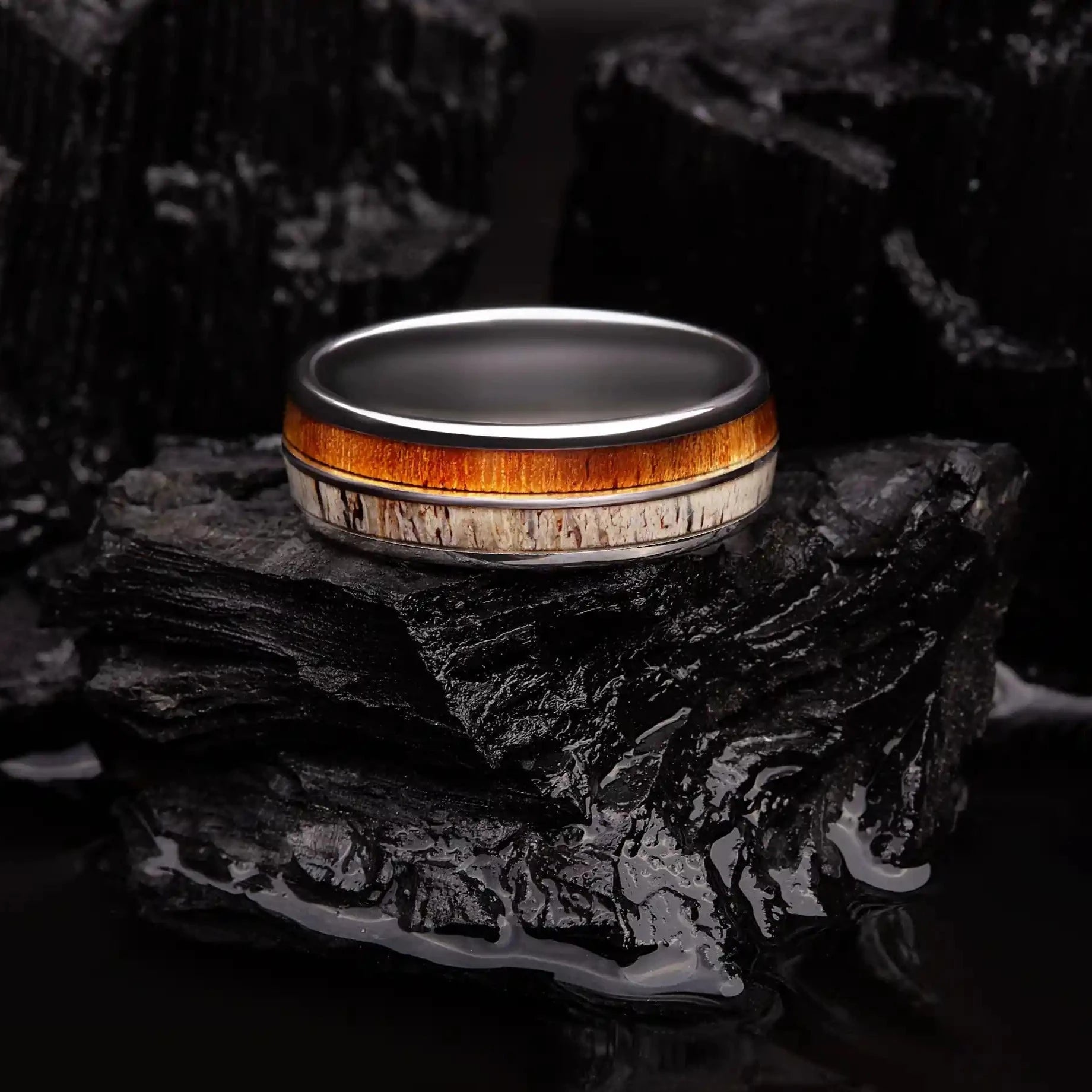 Silver brushed tungsten ring with deer antler and whiskey barrel oak wood. The ring is laying on a black coal