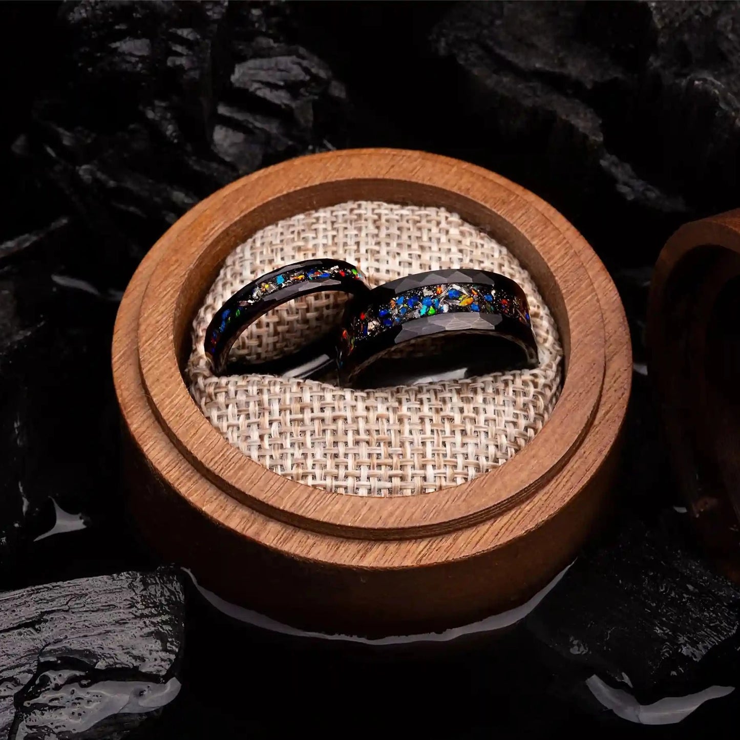 Two matching rings made of black hammered tungsten with blue opal and meteorite. The rings are placed inside a round wooden box.