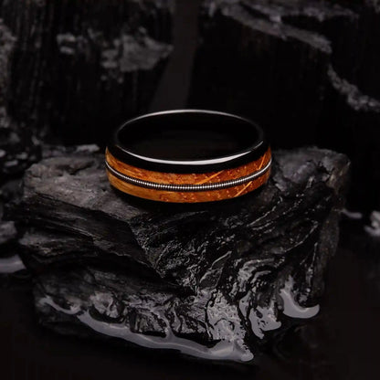 Black brushed tungsten ring with a guitar string and a whiskey barrel oak wood. The ring is laying on a black coal