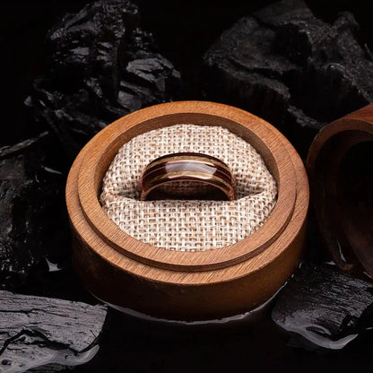 Brown brushed tungsten ring with guitar string and whiskey barrel oak wood. The ring is placed inside a round wooden box.